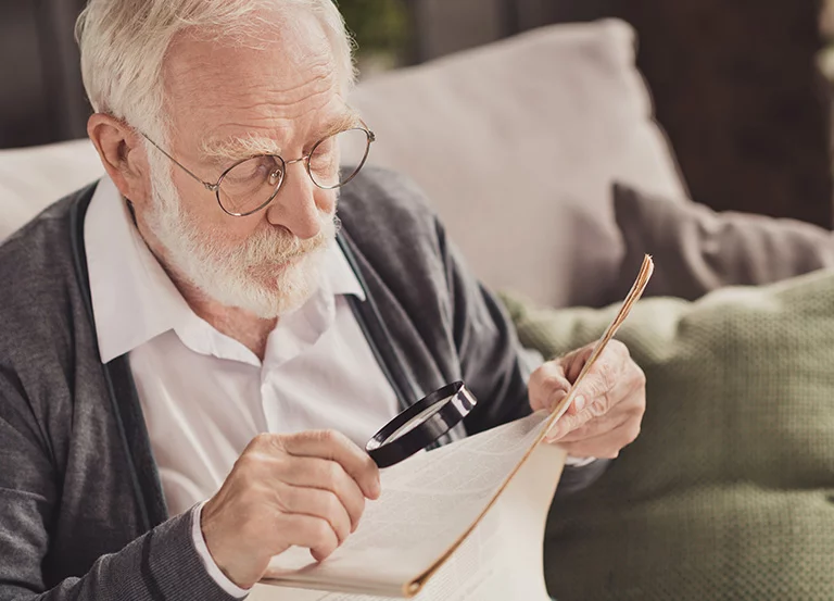 Starszy mężczyzna w okularach czyta gazetę przy pomocy lupy